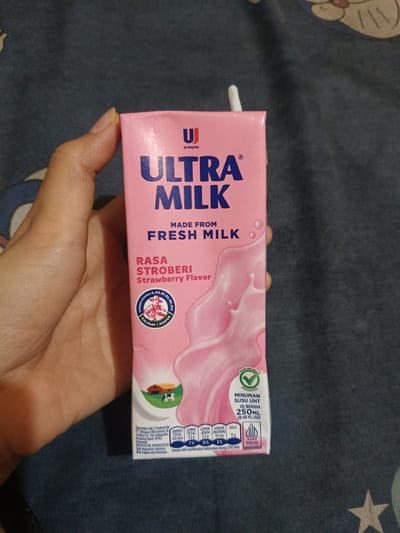 A light-skinned hand, with visible knuckles and short fingernails, grasps a 250ml pink Ultra Milk carton, strawberry flavor, on a dark patterned fabric background. A white plastic straw protrudes from the top right of the carton. The front of the carton displays "ULTRAJAYA" and "ULTRA MILK" in prominent blue lettering, with "MADE FROM FRESH MILK" and "RASA STROBERI / Strawberry Flavor" below. A graphic of swirling pink liquid suggestive of milk fills the lower right portion of the carton. A circular emblem on the left indicates "SUMBER VITAMIN A, D3, B1, B2, B6, B12 KALSIUM FOSFOR". At the bottom, a small image depicts a red barn and a white cow. Nutritional information lists "ENERGI 190 kkal", "LEMAK TOTAL 4.5g", "LEMAK JENUH 2.5g", "GARAM (NATRIUM) 50 mg", and "GULA 12 g", with corresponding daily value percentages. Other text identifies the product as "MINUMAN SUSU UHT", "ISI BERSIH 250ML (8.45 FL.OZ)", and includes a "HALAL INDONESIA" certification logo and a green "PILIHAN LEBIH SEHAT" (Healthier Choice) tick. The manufacturer is listed as PT. Ultrajaya Milk Industry & Trading Co. Tbk. in Kabupaten Bandung Barat 40973, Indonesia. The background consists of a dark blue-grey textile, likely a bedsheet, featuring faint, light-colored patterns, including a partial five-pointed star in the bottom right corner. The scene is set in the city of Cileungsi, Indonesia.