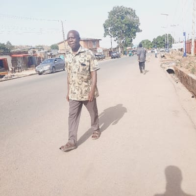 The image shows a man walking down a paved street in the city of Kufang, Nigeria. The man is wearing a tan shirt with a multi-colored design, brown pants, and brown sandals. He is walking towards the right side of the image. There are several buildings on the left side of the street, including a car dealership and a few shops. On the right side of the street is a sidewalk with a drain. A second man in a blue shirt is walking away from the camera in the distance. The sky is clear and there are no trees in the immediate foreground of the image. The photo was taken in Kufang, Nigeria. The latitude and longitude of the location are 9.863 and 8.867, respectively.