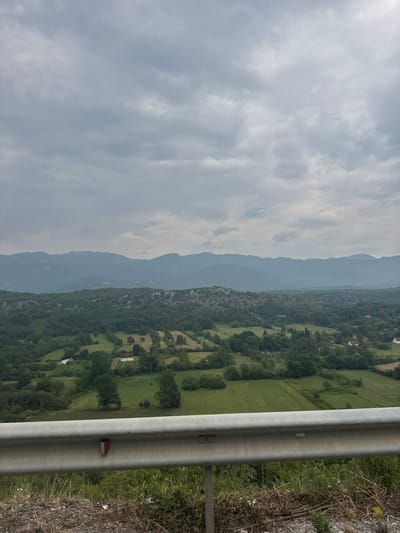 The image shows a scenic view of a rural landscape with rolling hills covered in lush green trees and fields. In the foreground, there is a metal guardrail, which appears to be part of a road or an overlook. The sky is overcast with heavy clouds, suggesting a potentially stormy or moody atmosphere. In the distance, a range of mountains is visible, fading into a hazy blue silhouette. The presence of scattered houses and cultivated fields indicates a settled rural area, possibly a village or small town. The overall impression is one of natural beauty and tranquility, characteristic of a mountainous region. The image does not depict any people or specific actions, focusing solely on the natural environment and a glimpse of human habitation. It is not possible to determine the exact location within the city of Frutak, Montenegro, from the image alone.