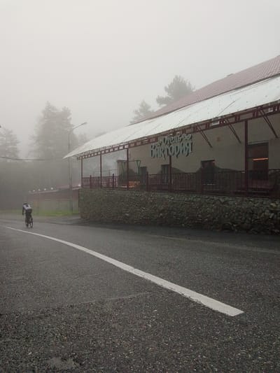 A lone cyclist, viewed from the rear, rides on a paved road on a foggy day. The cyclist is wearing a white and blue cycling jersey and black shorts, and is on a dark bicycle. The road has a white line dividing the lanes. To the right of the road is a building with a red-tiled roof and a covered outdoor seating area. A sign above the covered area reads "ОТЕЛЬ ВИКТОРИЯ" (Hotel Victoria). The building is set back from the road and is surrounded by trees, which are partially obscured by the fog. The overall environment is damp and overcast due to the fog.