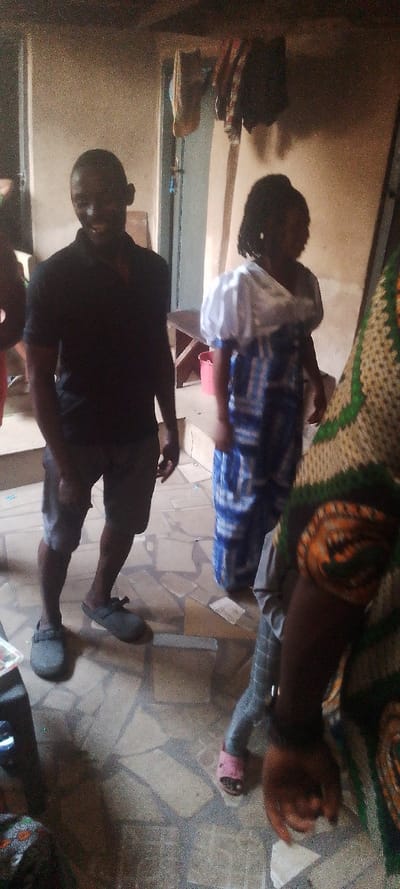 Inside a room with tiled floors, a man in a black shirt and shorts stands with gray sandals. To his right, a woman wears a blue and white dress with a white shawl. Behind them is a doorway and some clothes hanging. In the foreground, parts of another person wearing a patterned garment are visible. The lighting is somewhat dim.