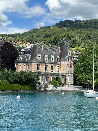 The image depicts a large, ornate building with a red brick facade and a dark, steep-pitched roof with multiple dormer windows. The building is situated on the shores of a bright blue body of water, likely a lake, with ripples on its surface. A white sailboat is docked to the right of the frame. In the background, a lush green, tree-covered hillside rises steeply, dotted with residential houses. The sky above is blue with scattered white clouds. The building's architecture suggests it might be a historic mansion or a government building, and it is decorated with flags at its entrance. The overall scene conveys a sense of tranquility and picturesque natural beauty. This scene is the exterior of the Schlössli Oberhofen, a castle located in Oberhofen am Thunersee, Switzerland.