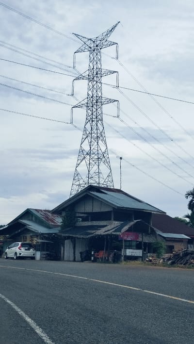 A vertical, eye-level shot captures a towering electricity transmission pylon against an overcast sky, with a cluster of buildings and a paved road in the foreground. The large lattice-style power tower, adorned with multiple power lines extending horizontally across the frame, dominates the upper and central portions of the image, silhouetted against the light grey, cloud-filled sky.
Below the massive pylon, a collection of rural or semi-rural structures line what appears to be a roadside. To the left, a white hatchback car is parked in front of a building with a corrugated metal roof. Adjacent to it, a central building features a steep, dark roof and an open-fronted section covered by a thatched canopy, under which chairs and an indistinct sign are visible; the sign appears to say "OPEN" and potentially some Indonesian text like "24 JAM". To the right, another building with a reddish roof stands, with a stack of firewood or logs nearby.
The foreground consists of a paved asphalt road, featuring a solid white line along its left edge and a solid yellow line marking the center. The diffused, natural light indicates daytime under a heavily cloudy sky. No individuals are present in the frame. The scene portrays a roadside environment in a residential or commercial area, consistent with the Lambarotunong, Indonesia context.