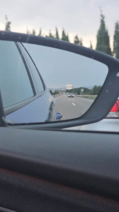 The image is taken from inside a vehicle, looking into the side-view mirror. The mirror reflects a highway with a few cars driving. The sky is overcast. The location is a highway outside of Calatorao, Spain, as indicated by the presence of a road sign with "Calatorao" written on it. The car in the foreground is dark blue. Another car, a silver sedan, is visible next to the camera car. The mirror shows a black car driving in the distance on the highway.