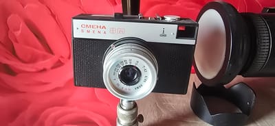 An eye-level, medium shot captures a vintage Smena 8M camera mounted on a tripod against a vibrant red, rose-patterned background. The camera, predominantly black with a silver top plate, is positioned in the foreground, its lens prominently featuring markings like "T-43 4/40" and various aperture and ASA settings. To the right of the camera, a large, cylindrical object with a white diffuser and a black lens hood is visible. The overall scene suggests a still-life arrangement, possibly in a home setting. There are no people or discernible actions taking place within the frame. The background's soft focus and the lighting emphasize the camera as the central subject. The location of Gostagayevskaya, Russia, is not visually represented in the image itself and can only be inferred from external information.