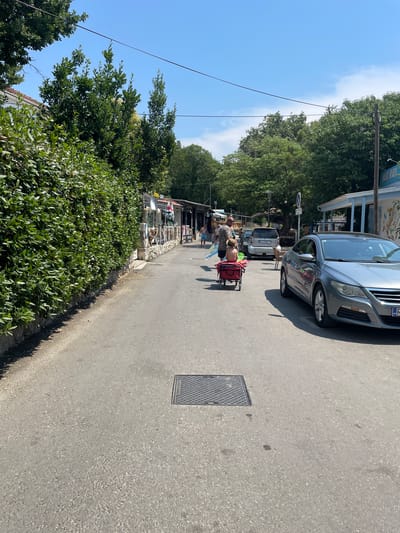 The image shows a sunny day in Crikvenica, Croatia. The scene depicts a street lined with shops and lush green trees. In the foreground, a person is seen with a red trolley and a beach umbrella, suggesting a leisure outing. A silver car is parked on the street, and another smaller car is visible behind it. The street appears to be paved, with a manhole cover visible on the right side of the road. The overall atmosphere is bright and relaxed, indicative of a pleasant day in a coastal town. Sunlight casts shadows on the pavement, hinting at the time of day, likely midday. Some text is visible on signs in front of the shops, but it is not clear enough to read. The presence of greenery and the style of the buildings suggest a Mediterranean setting.