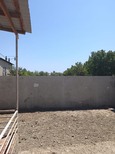 This image depicts an outdoor scene in Kasri Arifan, Uzbekistan. In the foreground, a gravelly yard is visible, with a section of a white metal structure on the left, possibly a pen or enclosure. Behind the yard, a tall, rough-textured concrete wall runs horizontally across the frame. The wall is weathered and shows signs of patching and wear. Above the wall, the clear blue sky is visible, with a few sparse green trees and foliage peeking over the top. To the left of the wall, a portion of a building with a window is visible, and above that, the corrugated roof of an open-sided structure is supported by wooden beams. Laundry clips are attached to a clothesline strung between poles. The overall impression is of a rural or semi-rural residential or agricultural setting.
