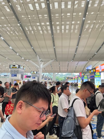 The image captures a crowded scene inside what appears to be a modern transportation hub, likely a train station or airport, in Longhua District, China. The ceiling is high and modern, with a patterned metallic structure and recessed lighting. A large crowd of people, predominantly adults, are seen standing and walking in various directions. Many are carrying luggage, such as backpacks and bags. People are dressed in casual attire. Some individuals are looking at their mobile phones. In the background, numerous digital display boards with colorful advertisements and information are visible, indicating a commercial and informational environment. The architecture suggests a contemporary public space designed to accommodate a large volume of travelers. The overall impression is one of a busy and active transit point.