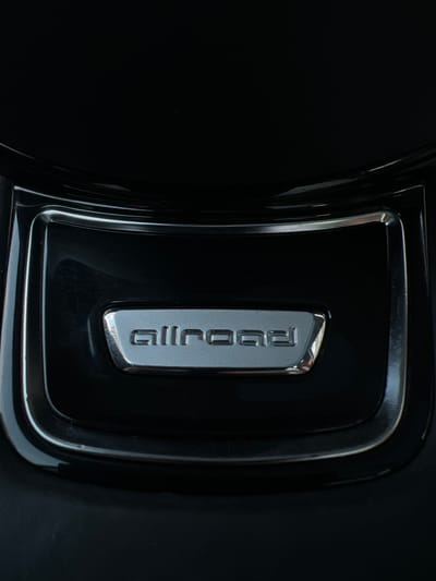 The image is a close-up, low-angle shot of a silver "allroad" emblem set into a black, glossy surface. The emblem is rectangular with rounded corners and features raised, sans-serif lettering. The surface surrounding the emblem has a curved indentation and appears to be part of an automobile's interior or exterior trim. The lighting suggests a somewhat reflective environment, with highlights visible on the glossy black surface and the metallic emblem. There are faint scuff marks on the black surface. There are no people or animals visible in the image. The location in Florești, Romania, is not determinable from the visual information provided.