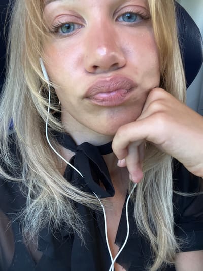 The image is a close-up, overhead shot of a woman's face and upper torso. She has blonde hair, blue eyes, and her lips are pursed. She is wearing white earbuds and a black top with a bow around her neck. Her right hand is resting on her chin, and her left hand is holding her hair near her ear. The background is dark, with hints of blue. The image appears to be taken in natural light. No specific location in Dorfen, Germany, is discernible from the image itself.