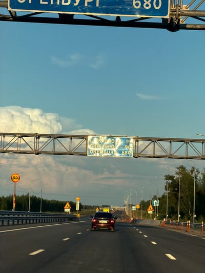 The image depicts a highway scene in Verkhnie Karmachi, Russia, likely during the daytime given the bright sky. The perspective is from a vehicle on the road, looking forward. The road is paved and has clear lane markings. To the left, a large blue signpost with white lettering is partially visible. The visible text on this sign is "680" and some illegible Cyrillic characters. Above the road, a metal framework supports another sign. This sign has yellow and blue sections and displays text in Cyrillic, including "190" and "680," along with "LAST CHISTOPO" and "ORENBURG."
Along the right side of the road, there are several traffic signs. One prominent yellow triangular sign shows a person walking. Another circular sign indicates a speed limit of "50." Traffic cones are also visible along the roadside, suggesting possible road work or a temporary lane closure. A dark-colored car is driving ahead on the highway. The overall scene is one of travel on a rural or suburban highway, with typical road infrastructure and signage. The sky is blue with some white clouds, indicating fair weather.