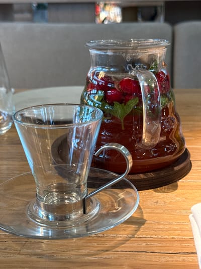 A glass pitcher filled with a red beverage, cherries, and mint sits on a dark wooden coaster on a wooden table. Next to the pitcher is an empty glass tea cup with a metal handle, resting on a clear glass saucer. The background shows a blurred seating area with upholstered furniture. The scene appears to be indoors at a restaurant or cafe located in Агаларов Эстейт Кп, Russia.