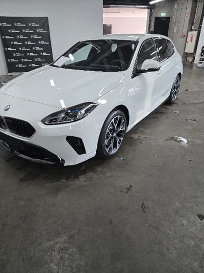 The scene is an interior view within a commercial facility located in Sprendlingen, Germany. A white, compact BMW hatchback is centrally positioned, oriented diagonally with its front towards the left foreground. The vehicle features clean white paintwork, modern headlamps, a black kidney grille, and multi-spoke dark alloy wheels. A white document is visible on the passenger side of the windshield. In the left background, a black banner with repeated "BMW MOTOWORLD" branding in white text and logos is displayed against a light-colored wall. The rear background includes an industrial ceiling with overhead lighting, a dark door, vertical conduits, and a red fire safety cabinet mounted on a light-colored wall. The floor is dark grey concrete, marked with several wet patches and smudges, predominantly in the foreground and near the car's front right wheel. No individuals are present, and no actions or interactions are depicted.