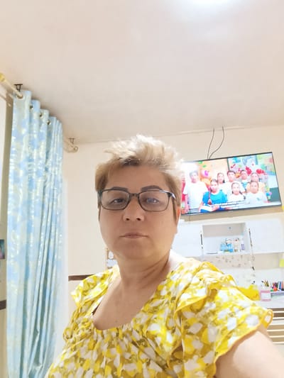 This media file is a selfie taken by a woman with short, blonde hair and glasses. She is wearing a yellow and white floral patterned top. In the background, a large flat-screen television displays a group of children, possibly from a TV show or movie. To the left of the woman, there are light blue curtains with a white pattern. Below the television, a white shelf unit is visible, containing various items including what appears to be figurines and art supplies. The scene appears to be indoors, likely in a residential setting. The lighting suggests it could be daytime. No specific emotions are overtly conveyed. There is no visible text in the image.