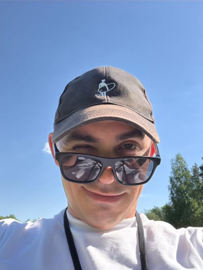 A medium shot captures a person wearing sunglasses and a cap against a backdrop of blue sky and distant greenery. The person is positioned in the foreground. The city of Oreshniki, Russia cannot be determined from this image.