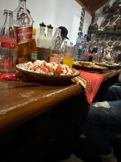 An indoor dining scene in Terziysko, Bulgaria, features a wooden table laden with food and beverages, suggesting a social gathering. In the immediate foreground, a dark ceramic bowl holds a salad composed of diced red tomatoes, green components, and shredded white cheese. To the left of the salad, an uncapped Johnnie Walker Red Label whisky bottle, partially full, stands next to an empty glass Coca-Cola bottle. Further back, several other empty clear glass bottles, a small orange condiment bottle, and a glass containing a yellow liquid are present. A woven textile with red, black, yellow, and green stripes serves as a table runner, partially draped over the table edge to the right. The wooden table surface exhibits signs of wear.

In the mid-ground, additional plates of food, various beverage bottles including a "DEVIN" branded water bottle, and multiple glasses are arranged along the table. Several individuals are partially visible and out of focus in the background, seated around the table. One person is identifiable by a black baseball cap worn backward. The lower right foreground shows the upper thigh of an individual wearing dark denim jeans with a noticeable tear or distressed patch. The background includes a white wall and a stone-clad wall adorned with pottery items, beneath a wooden ceiling beam.