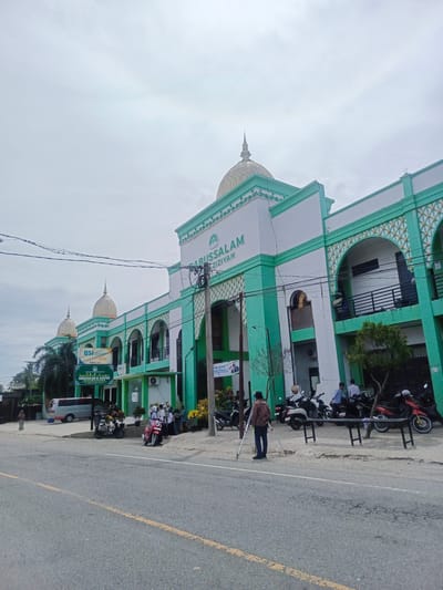 The image shows the front of a large, two-story building with a distinctive green and white facade, featuring multiple arched windows and several domes topped with golden spires. The building is identified by a sign as "BABUSSALAM AL-AZIZIYAH." Adjacent to the building, a road runs horizontally across the foreground. Several motorcycles are parked along the side of the road, and a group of people are gathered near the entrance of the building. A white van is parked further down the road. The sky is overcast. The scene appears to be in a public area, possibly a town or city. There are power lines visible overhead.