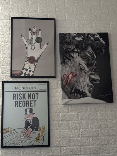 The image displays an indoor scene in the city of Breda, Netherlands, featuring three distinct artworks mounted on a white, textured wall with a brick-like pattern.
The top-left artwork is a vertically oriented, black-framed print depicting a stylized human hand with pale skin and black fingernails, adorned with various rings (including a butterfly, a leopard head, a green gemstone, and a red gemstone) and a pearl necklace. The background is light gray with a black and white checkered pattern at the bottom, and a small, partially obscured white tag is visible in the lower-left corner.
To the right of this, the top-right artwork is a vertically oriented canvas print portraying a grayscale, profile view of Michelangelo's David bust. The bust is overlaid with graffiti-style elements, including black text "AXA" on the forehead, a red outlined heart on the cheek, black text "Peace" on the side of the face, and dripping black text "Love" below the chin. A white signature, "Chris Bellini," is present in the lower right.
The third artwork, positioned below the hand print, is a vertically oriented, black-framed print designed like a newspaper front page. It is titled "WEEKLY IKONICK NEWS" and "MONOPOLY," with subtitles "OFFICIAL ISSUE" and "EST. 1935." The prominent headline reads "RISK NOT REGRET." The illustration depicts the Monopoly Man, in his iconic top hat and suit, leaning over a craps table, actively rolling two dice. Stacks of gold coins are arranged on both sides of the table.
The artworks are arranged in a staggered formation on the wall. No individuals are present in the depicted environment.