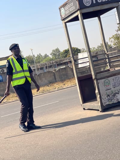 This image displays a daytime scene on an asphalt road in Basawa, Zaria, Nigeria. A male individual, identified as a police officer or traffic warden by his uniform, stands on the road towards the left of the frame. He wears a black beret, a black short-sleeved shirt with a bright green-yellow reflective vest, dark trousers, and dark dress shoes. He holds a dark, rigid baton vertically in his left hand, while his right arm is at his side. He looks towards the viewer.

Long, distinct shadows from the officer and other objects are cast onto the road, indicating bright, direct sunlight. To the right, a metallic, open-frame structure, likely a kiosk or shelter, stands on the roadside. Its top section features a sign reading "HEALTH FOR BASIC NEEDS." A sign on its lower right side advertises "Elite Intl College, Science &..." established 2018.

In the background, behind a concrete fence, a prominent white sign clearly states "NIGERIAN SCHOOL OF SOIL SCIENCE, BASSAWA ZARIA." Further behind, buildings with visible air conditioning units, utility poles, and distant green trees are present under a clear, blue sky.