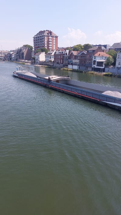 An aerial, medium shot shows a barge carrying gravel on the Sambre River in Namur, Belgium. The barge is positioned in the center of the frame, moving from left to right. Along the right bank of the river, a row of diverse buildings forms the cityscape. The buildings vary in architectural style and height, with some featuring red brick facades and pitched roofs, while others are more modern. A tall, reddish-brown building with multiple stories stands out among the others. The river is calm with ripples on the surface. The sky is clear and blue with a few white clouds. The overall scene depicts a tranquil urban waterway with industrial and residential elements.