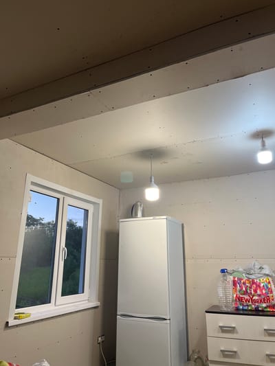 The image depicts an interior scene, likely a room under renovation, located in Клобушнево, Russia. The room is sparsely furnished, with an unfinished drywall ceiling and walls, evident from the exposed screw heads. Two modern pendant lights with white shades hang from the ceiling, illuminating the space. A white refrigerator stands against the right wall, and next to it, a black and white dresser with two drawers is visible, topped with a large clear plastic water bottle and a colorful tote bag from "New Yorker". A window with white framing is on the left side of the room, offering a view of green trees and the sky outside. The overall impression is one of a functional space in transition, possibly a kitchen or utility area. There are no people or animals in the image.