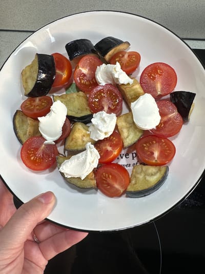 A human hand, with fair skin and a thin silver-colored ring on the left ring finger, holds a white round ceramic plate with a thin black rim from the lower-left side. The plate contains a casual arrangement of cooked food.

Numerous pieces of eggplant, appearing as golden-brown wedges with their dark purple skin visible on the outer edge, are scattered across the plate. Interspersed among the eggplant are multiple halves of bright red cherry tomatoes, exposing their seeds and juicy interiors. Several distinct dollops of a thick, opaque white creamy substance, consistent with soft cheese or a dairy product, rest on top of various eggplant and tomato pieces. Faint, partially obscured light grey text, possibly reading "ve p" and "res of su," is visible on the plate's surface beneath the food.

The background above the plate features a light grey, mottled or textured surface, likely a wall or countertop. Below and to the right of the plate, a dark, reflective surface, possibly a kitchen stovetop with a subtle white curved marking, is visible. The scene is brightly and evenly lit. The image contains no identifiable features or contextual clues linking it to Monchegorsk, Russia, or any other specific geographical location.