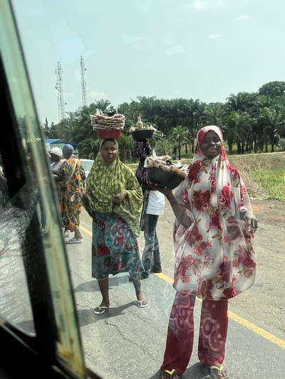 The image captures a vibrant roadside scene, likely viewed from inside a moving vehicle given the visible window frame and reflections in the foreground. The primary subjects are several individuals, predominantly women, on what appears to be an asphalt road.
In the mid-ground, a woman in a light green patterned top and a colorful patterned skirt carries a reddish pot or basket, filled with stacked items resembling dried fish or food, balanced on her head. She wears white flip-flops and looks directly towards the viewer with a neutral expression. Beside her, another woman in a light-colored floral head covering and matching loose-fitting floral top over dark red patterned trousers stands, also facing the camera with a faint smile. She wears yellow flip-flops. Behind these figures, other people, including what appears to be a child in an orange and black patterned outfit, are partially visible.
The setting is characterized by a bright, sunny day with a clear sky. Palm trees line the background, suggesting a tropical or sub-tropical environment. Two tall metal structures, possibly communication towers or electricity pylons, are visible against the sky in the distance. A yellow line is painted on the road surface.
The activity taking place appears to be roadside commerce, with individuals possibly hawking goods to passing vehicles, a common sight in Nigeria. There is no discernible text visible in the image. The overall impression is one of everyday life along a rural or semi-rural road in Nigeria.