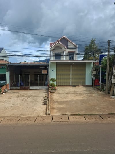 The image captures a street-level view of two adjacent buildings in Tân Phú 2, Vietnam. The building on the left has a green facade and appears to be a shop, with a glass door and a car visible inside. A sign with Vietnamese text, including "HUA XE," is displayed. The building on the right is lighter in color with a closed, corrugated metal gate. Above the right building, a two-story house with a red-tiled roof is visible. Overhead, a dense network of electrical wires crisscrosses the sky, which is overcast with dark clouds. The ground in front of the buildings is a concrete or asphalt surface, with a concrete drainage channel running along the edge of the road. A potted plant with pink flowers sits near the right building. The overall scene depicts a typical urban streetscape with commercial and residential structures.