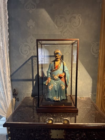 A doll-like figure encased in glass sits on a dark, elaborately carved wooden box or chest. The figure wears a pale blue robe decorated with floral patterns, and holds an object in its hands. A stripe of blue paint marks its forehead. The background consists of a wall decorated with a stylized, repeating pattern, and a white lace curtain is visible to the left. A wooden panel or curtain hangs on the right. The scene is bathed in natural light, with strong shadows cast by the glass enclosure. The setting is likely a room within the Palácio Nacional de Sintra, Sintra, Portugal.