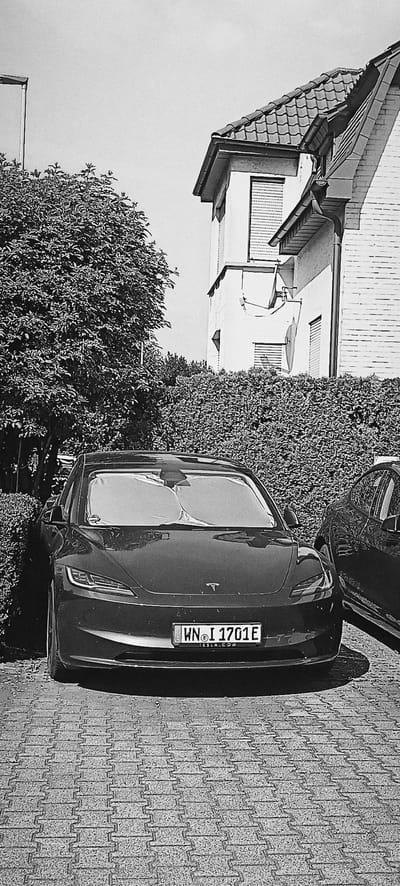 A black Tesla Model 3 is parked on a cobblestone driveway in front of a building with a pitched tile roof. The car's license plate reads "WN I 1701E". A second black car is partially visible to the right of the Tesla. A dense green hedge forms a backdrop for the cars, with a large tree to the left of the Tesla. The building appears to be a residential property, with a satellite dish mounted on its exterior wall. The image is rendered in black and white and has a grainy texture. The location is identified as Endersbach, Germany.