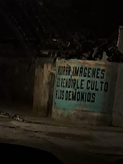 A close-up, dimly lit, eye-level shot captures graffiti on a weathered, turquoise surface. The text, rendered in bold, black capital letters, reads: "ADORAR IMAGENES ES RENDIRLE CULTO A LOS DEMONIOS." The graffiti is partially obscured by shadows and appears to be located outdoors, possibly on a wall or structure within the city of Cumana, Venezuela. The surrounding environment is dark and indistinct, with hints of rough textures and debris.
