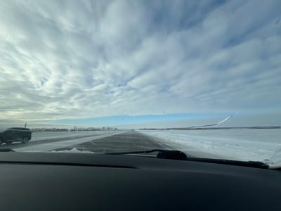 The image depicts an outdoor winter scene in Morskoi Chulek, Russia, viewed from inside a vehicle through its windshield. The dark dashboard and a windshield wiper are visible at the bottom of the frame. A dark, cleared paved surface, likely a road or runway, extends centrally into the distance, flanked on both sides by vast, flat fields covered in snow and ice. A dark-colored SUV or truck is partially visible on the left side, positioned parallel to the paved surface. Along the distant horizon, a subtle line of dark vegetation or trees is present. The sky transitions from a light blue near the horizon to a heavily clouded expanse of textured grey and white clouds overhead. A distinct, curvilinear white snowdrift or structure rises from the snowy ground on the far right horizon. Multiple small, dark specks are discernible in the mid-sky, possibly birds. The scene is illuminated by natural daylight under generally overcast conditions.