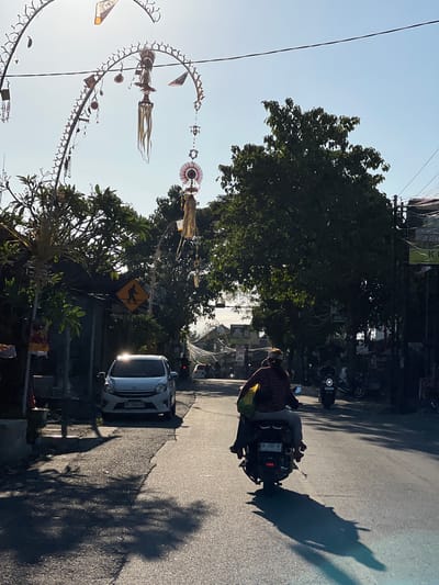 The image depicts a street scene in Kubu, Indonesia, bathed in the warm glow of the late afternoon sun. In the foreground, two individuals are riding on a motorcycle, viewed from behind. The rider wears a striped shirt and a head covering, while the passenger, positioned behind the rider, is seated. The motorcycle itself has a license plate visible, reading "DK 4740 NF".
The road surface is asphalt, showing signs of wear and tear. Shadows of trees and decorative arches are cast across the road, indicating the presence of vegetation and festive decorations. A white car is parked on the left side of the road, with its headlights reflecting the sun. Beside the car, a yellow pedestrian crossing sign is mounted on a pole.
Further down the street, other motorcycles and vehicles are visible, suggesting ongoing traffic. Buildings and trees line both sides of the road. Overhead, decorative arches, adorned with flags and streamers, span across the street, possibly part of a local celebration or cultural display. The sky is clear and blue, contributing to the bright, sunny atmosphere.