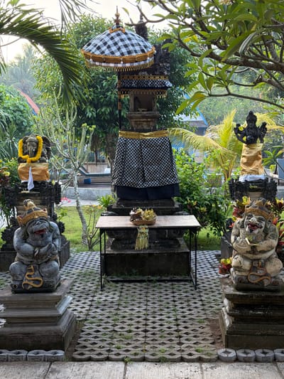 The image captures a courtyard in Toyapakeh, Indonesia. In the center stands a small Balinese shrine, draped in a black and white checkered cloth, with an ornate umbrella shading it. A table holds a small offering of leaves and flowers. Two stone guardian statues, decorated with gold accents, flank the entrance to the shrine. The ground is covered in interlocking concrete pavers. Lush tropical foliage surrounds the shrine, with glimpses of buildings in the background. The scene is captured on a slightly overcast day.