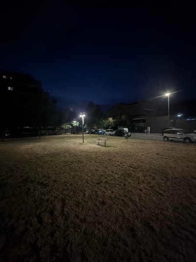 This is a nighttime photograph of a park area in Novara, Italy. A tall, dark building is visible in the background to the left, with illuminated windows. A street runs parallel to the park, lined with parked cars. Two streetlights illuminate the park, casting a warm glow on the dry, mown grass. A wooden bench is situated in the park, facing the street. The sky is a deep blue, indicating it is twilight or early night. The overall scene is calm and quiet, suggesting a residential area.