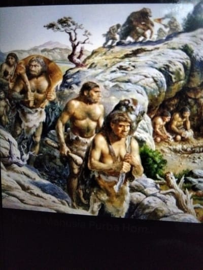 This is a painting depicting a prehistoric scene with several early humans in a rugged, rocky environment with sparse vegetation. In the foreground, a muscular man carries another person on his shoulders. To his right, another man stands, holding a wooden tool. In the background, several other individuals are visible, some gathered around a fire or engaged in activities within a cave-like structure. The landscape includes rocks, a solitary tree with twisted branches, and distant mountains under a hazy sky. The text at the bottom, partially obscured, reads "Ka... Manusia Purba hom...". This text suggests the painting might be related to early human history in Indonesia, possibly referencing "Manusia Purba" which translates to "ancient humans." However, there is no specific indication of the city of Kualaraja, Indonesia, within the visual content of the painting itself.