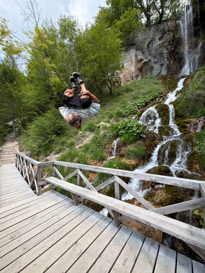 The image shows a man seated near a waterfall and a wooden bridge. He is wearing a light-colored, tie-dye shirt, black shorts, and black shoes. He sits with his knees drawn up to his chest, located amidst lush green trees and vegetation. To the right, a rustic wooden bridge stretches across the scene, its planks showing visible grain. The waterfall cascades over a rocky cliff covered in moss and greenery. The photo was taken at the Sopotnica Waterfalls, located in Prijepolje Municipality, Serbia. Wooden stairs are partially visible.