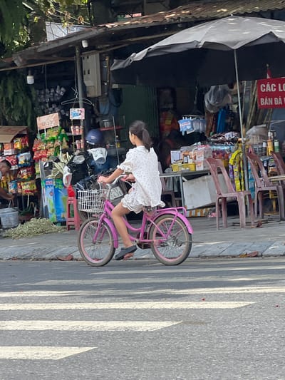 A young girl with dark hair pulled back in a ponytail, wearing a white floral dress, rides a pink bicycle with a white basket on a street in Tân Quang Commune, Vietnam. The girl is facing away from the viewer and to the left, her feet on the pedals. The bicycle has pink rims. The background shows a bustling street scene with a covered market stall selling various goods, including snacks and drinks. A motorcycle is parked behind the stall. Several plastic chairs are visible in front of the stall. A red sign with white text, appearing to be Vietnamese, is partially visible to the right. The street has a pedestrian crossing marked with white stripes. The lighting suggests it is daytime, with some shadows cast on the road.