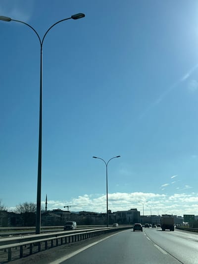 The image presents a daytime scene on a multi-lane highway in Sancaktepe, Turkey, viewed from a vehicle looking forward. A tall, dark grey streetlight pole with a bifurcated arm supporting two light fixtures is prominent in the left foreground, silhouetted against a bright blue sky. A metal guardrail runs along the left edge of the highway. The asphalt road recedes into the distance, accommodating several vehicles traveling away from the viewer, including a white commercial van in the left lane, a dark sedan, and a large tan dump truck in the right lane. Other vehicles are visible further down the road, along with additional streetlights. The background features an urban environment with numerous buildings of varying heights, residential structures, and bare deciduous trees. A slender mosque minaret is visible to the left of the highway in the mid-distance. A large yellow construction crane extends across the skyline, indicating active development. The sky is predominantly clear blue, with scattered white cumulus clouds near the horizon and a sun flare visible in the upper right quadrant. No individuals are distinctly identifiable.