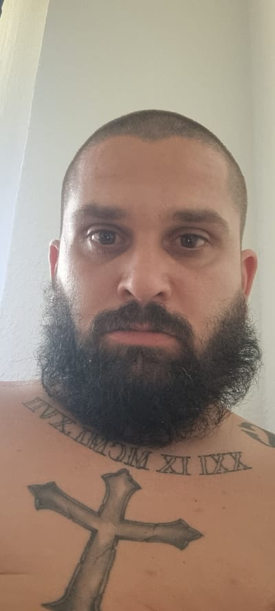This media file is a close-up, low-angle portrait of a man. The man has a shaved head with a short beard and appears to be looking directly at the camera with a neutral expression. Visible on his chest is a tattoo of a cross, and next to it, a tattoo of Roman numerals that appear to read "XXIX MCM." The lighting suggests an indoor setting with soft, diffused light. The background is a plain, light-colored wall. The rotation of the image indicates it may have been taken with a mobile phone held in a portrait orientation and then rotated. There are no discernible environmental cues to suggest the specific location within Mehltheuer, Germany, other than the general indoor setting.