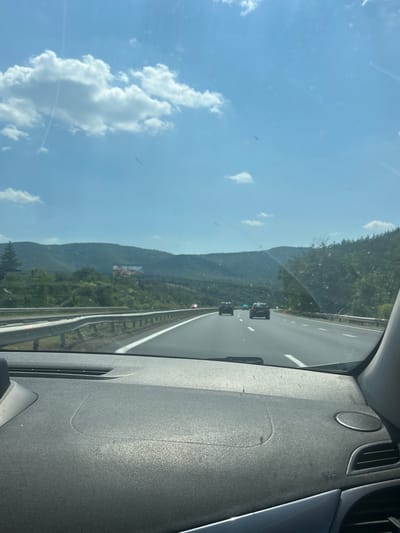 A view from the inside of a vehicle traveling on a highway in Dolno Varshilo, Bulgaria. The highway stretches forward, flanked by guardrails and green, hilly landscapes under a bright blue sky with scattered white clouds. Several cars are visible on the road ahead, and a billboard can be seen on the hillside in the distance. The dashboard of the vehicle is in the foreground.