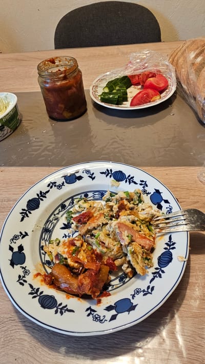 The image shows a top-down view of a meal laid out on a wooden table. The primary subject is a plate of scrambled eggs, mixed with what appears to be pieces of sausage or ham, and garnished with chopped green onions. The eggs are served with a portion of what looks like a tomato-based sauce or relish. A fork is placed on the plate, indicating the food is ready to be eaten.
To the left of the plate, there is a small white dish containing sliced tomatoes and cucumber. Adjacent to this dish is a glass jar, partially filled with a reddish, chunky substance, likely a condiment or spread. Further left, a dark, textured surface, possibly a chair or cushion, is visible. In the bottom right corner, part of a white container with visible text is peeking into the frame. The text appears to say "Frischkäse," which translates to "cream cheese" in German.
The overall setting suggests a casual meal, possibly breakfast or lunch, in a domestic environment. The lighting suggests it could be daytime, with shadows cast on the table. There are no people visible in the image. The scene is set in Oelsberg, Germany.
