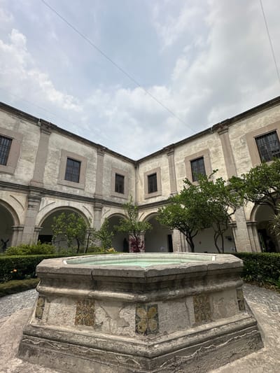 The image depicts a tranquil outdoor courtyard of a historical building, likely in Tepotzotlán, Mexico, under an overcast or partly cloudy sky during daytime. The primary subject is a multi-story structure with a weathered, light-colored stone facade accented by darker stone around rectangular windows and arched porticos. Each floor features multiple barred windows, and below them, open arches lead to covered pathways.
Within the courtyard, vibrant green trees with visible branches grow, their foliage providing a natural contrast against the aged stone architecture. In the foreground, positioned to the right, stands a substantial stone structure, possibly a fountain or a large planter. This structure is adorned with colorful ceramic tiles featuring intricate geometric and floral patterns, adding a decorative element to the robust stone. The tiled patterns are visible on the side and near the basin of the structure.
The scene conveys a sense of historical grandeur and peacefulness. No human activity or visible text is present. Thin lines or wires are subtly discernible against the light grey sky in the upper portion of the image. The overall impression is one of architectural beauty and natural integration within a historic setting.