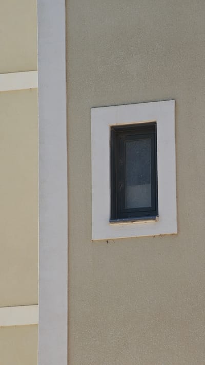 The image shows a small, rectangular window set into a beige, textured stucco wall. The window has a dark frame and appears to be made of tinted glass. A white, rectangular trim surrounds the window, creating a raised border. Below the window, there is a horizontal white band that runs across the image, separating the upper wall from a lower section that is a lighter shade of beige or yellow. This lower section is partially obscured, with what appears to be a white vertical element visible in the bottom center of the frame, suggesting a structural feature or architectural detail. The lighting suggests it is daytime, and the overall impression is of an exterior architectural detail. The scene is devoid of people or any activity, focusing solely on the building's facade. The location context provided is Alhama De Aragón, Spain, which suggests this is likely an outdoor shot of a building in that area. There is no visible text in the image.