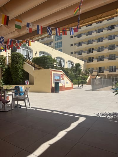 The image depicts an outdoor courtyard at the Hofmann Hotel in Fuengirola, Spain, bathed in bright sunlight. A row of national flags hangs across the top of the frame, strung from a beige awning. The courtyard itself is paved with large, square tiles. In the background, a multi-story hotel building with cream-colored walls and dark balconies dominates the scene. To the left, a yellow building with ivy-covered walls and an outdoor staircase leads up to the main hotel structure. A glass-enclosed area, possibly a pool enclosure, is visible on the right. In the foreground, a table with chairs suggests an outdoor dining or seating area. One chair has a blue towel draped over it. A person's legs and feet, wearing flip-flops, are visible at the lower left of the frame, seated at the table. Shadows from the awning and foliage are cast across the courtyard, indicating a sunny day.