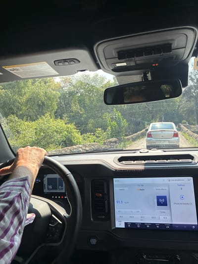 The image is taken from inside a vehicle, presumably a Ford Bronco, showing the dashboard and the driver's perspective. The driver's hand is visible on the steering wheel, wearing a purple and white checkered shirt. The speedometer indicates 16 km/h, and the gear selector is in the 'Drive' position. The infotainment screen displays a radio station tuned to 91.3 FM. Through the windshield, a silver car is visible on what appears to be a narrow road lined with vegetation. The license plate on the car has some partially visible characters, including "HML-373-". It is daytime, and the weather seems clear. Based on the license plate style and the general environment, it is highly probable that the location is in Mexico, specifically Tepeji de Ocampo, as the image's source indicated.