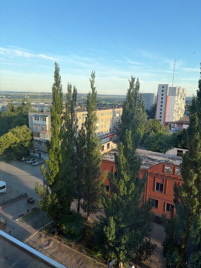The image depicts a view of an urban environment in Skopin, Russia, characterized by a mix of residential and commercial buildings, as well as significant tree cover. The scene is captured under a clear blue sky with a few wispy clouds.

In the foreground, tall, slender trees with dense green foliage dominate the view. To the left, a white van is parked on a paved area next to a building with a brick lower section and a lighter upper facade. Several cars are parked in a row further down the street.

Mid-ground structures include multi-story apartment buildings, some with balconies and visible windows. One prominent building is white with orange accents and a tall antenna on its roof. Adjacent to it is a shorter, light-colored building with multiple windows. There's also a brick building with numerous dark-framed windows visible in the middle right of the frame. A flatter, red brick building with open window frames is also visible in the lower right portion of the image.

The background reveals a more distant urban landscape with more buildings and trees stretching towards the horizon, suggesting a moderately sized city. The overall impression is a daytime scene, likely in the late afternoon or early morning due to the angle of the sunlight casting shadows and illuminating the scene. No people are visible in the image. The environment appears to be a well-established urban area with a notable presence of mature trees interspersed among the architecture.