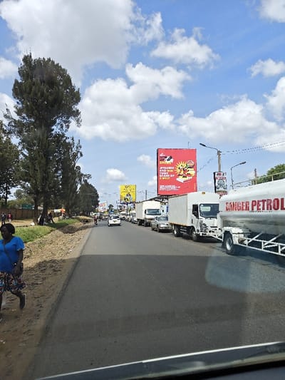A daytime street scene features a line of vehicles, including a large tanker truck labeled "DANGER PETROL," parked on the right side of a two-lane road. A woman walks along the roadside on the left. Billboards are visible in the background, one red and one yellow. The sky is partly cloudy. The image appears to be taken from inside a vehicle.