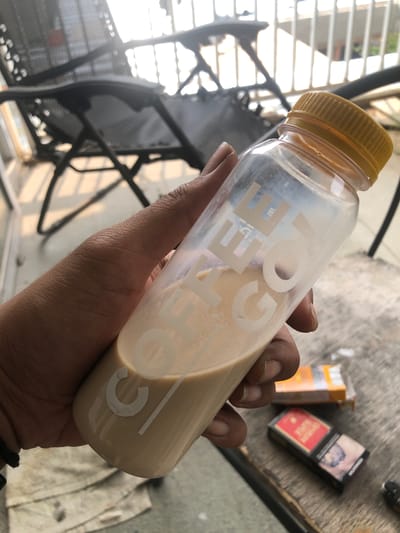A close-up shot captures a person's hand holding a clear plastic bottle filled with a light brown, possibly coffee-based, beverage. The bottle has a yellow ribbed screw cap and features white, stylized text that spells "COFFEE" vertically along its side. Some froth or settled particles are visible within the liquid. The hand holding the bottle appears to have dark skin, and the fingers show unmanicured nails.
In the blurred background, two cigarette packs are visible on a textured, grey surface, likely a table or floor. One pack is predominantly red with white lettering that clearly reads "DJARUM SUPER" and "PERINGATAN" (Indonesian for "WARNING"), indicating an Indonesian brand. Another yellow/orange cigarette pack is partially visible next to it, with text too blurry to decipher. Behind these items, there's a suggestion of a metal outdoor chair or railing and a white cloth.
The scene is brightly lit by natural light, suggesting daytime, and strong highlights and shadows are present, especially in the upper left background, hinting at an outdoor setting like a balcony or patio. The overall context, including the specific cigarette brands, places the scene in Cibinong, Indonesia.
