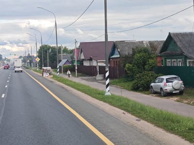 The image depicts a road in the city of Maryino, Russia, with houses lining the right side of the road. The road is paved and has a yellow line marking the lane. Several cars are visible on the road, including a silver SUV parked on the side. There are also streetlights and power lines overhead. The houses appear to be residential, with various architectural styles and colors. A person is walking on the sidewalk in front of the houses. There is also a market stall by the side of the road selling produce. The sky is overcast, with clouds visible. The overall scene suggests a suburban or rural environment.